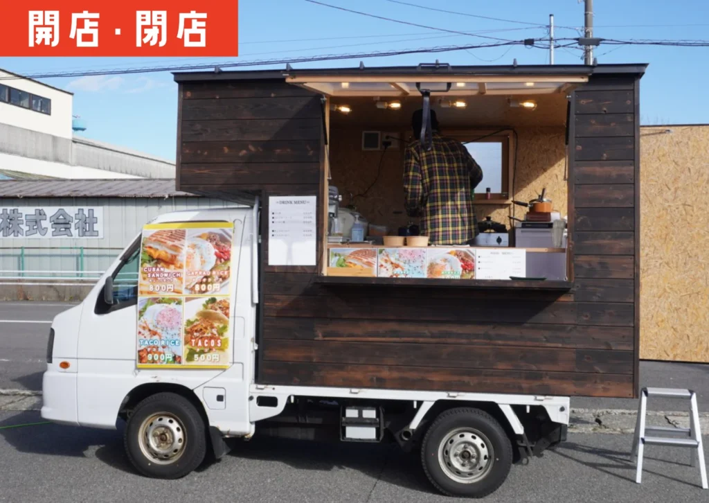 黒い木目調のキッチンカーが駐車場に停まり、側面の大きなメニュー写真にはキューバサンドやタコライスなどの料理が並んでいる。車内では店主と思われる人物が調理をしている様子。