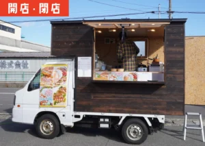 黒い木目調のキッチンカーが駐車場に停まり、側面の大きなメニュー写真にはキューバサンドやタコライスなどの料理が並んでいる。車内では店主と思われる人物が調理をしている様子。