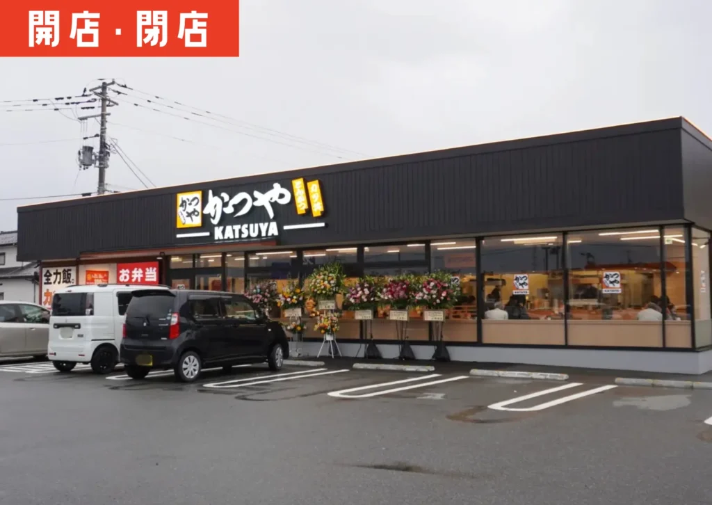 酒田市東大町にオープンしたとんかつ・カツ丼チェーン「かつや」店舗外観。黒を基調とした新店舗の前に開店祝いの花が並び、駐車場には多くの車が停まっている様子。